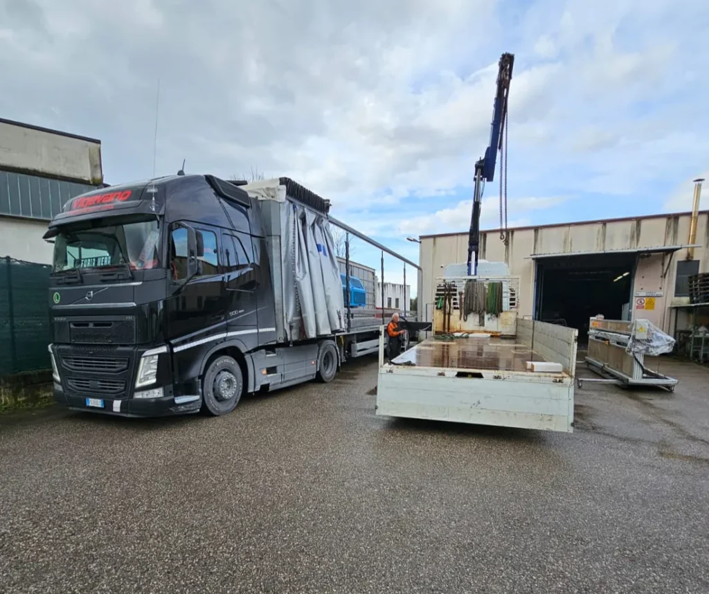 Camion con gru che scarica il carico su un mezzo a cassone nel piazzale di un capannone